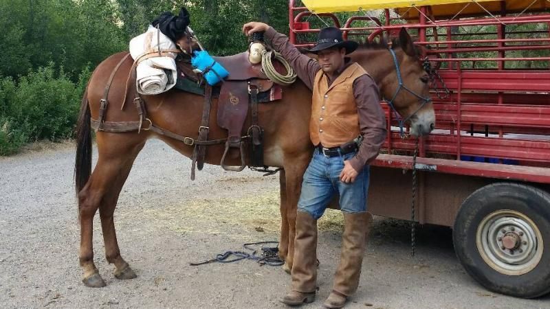 A person in western attire stands next to a saddled horse beside a red livestock trailer in a gravel lot.
