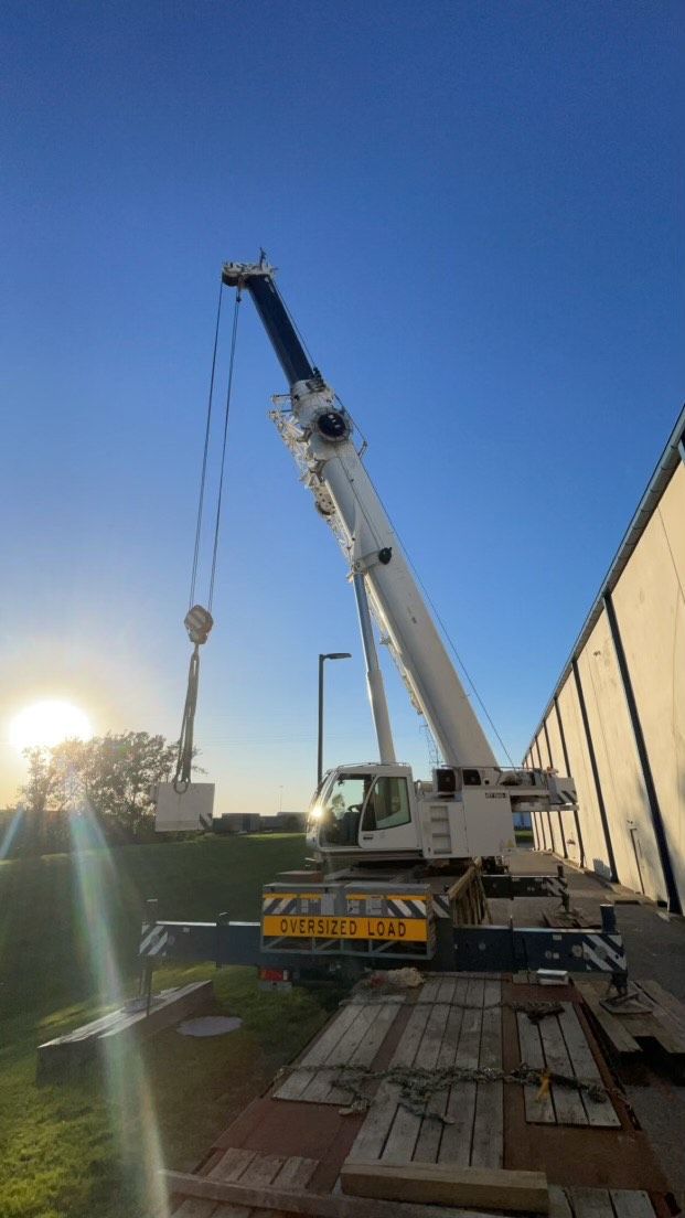 A white crane lifting an object at a construction site, with a bright blue sky and sunlight in the background.