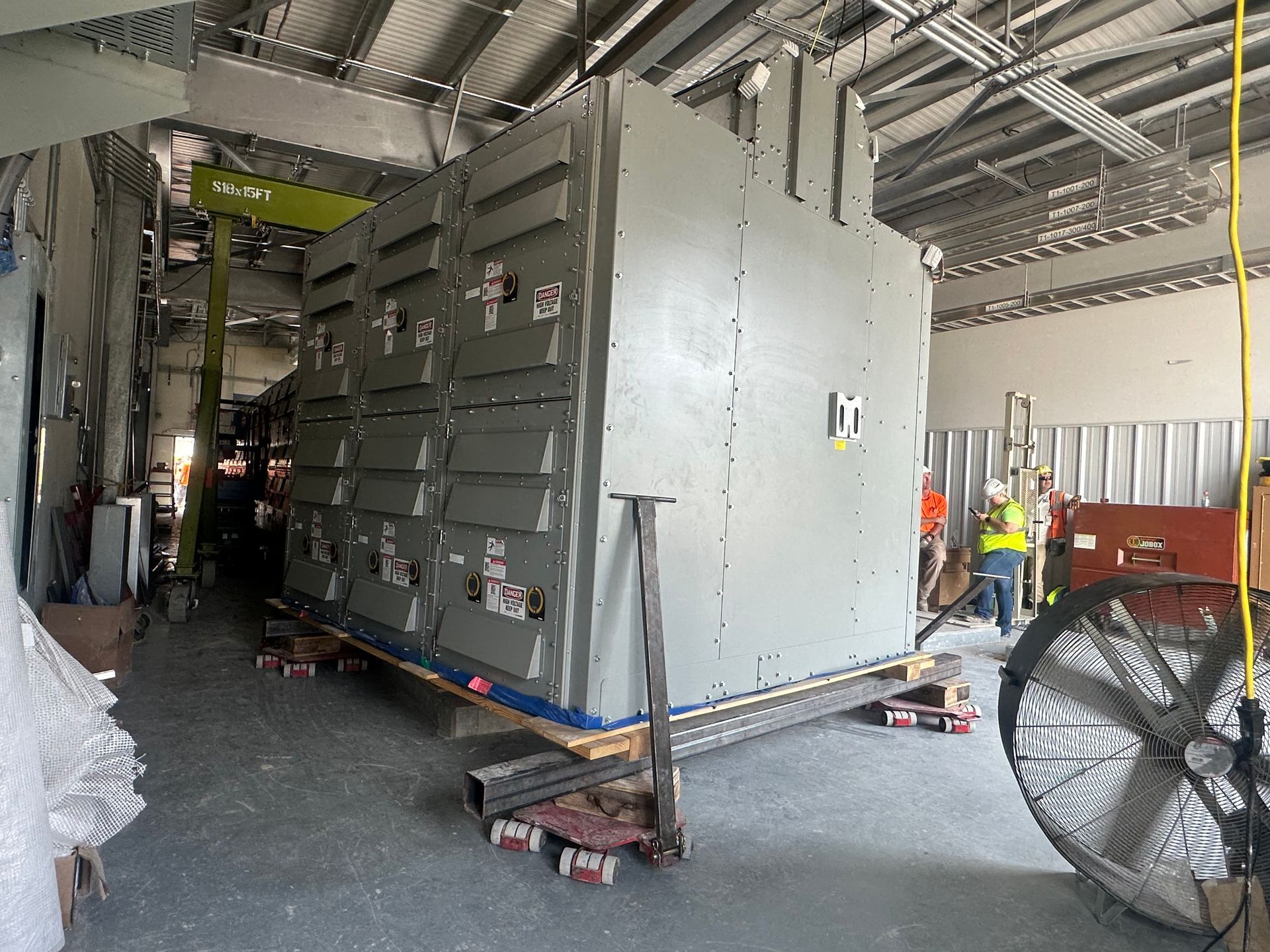 Large gray electrical equipment being moved in a construction site; workers in the background.