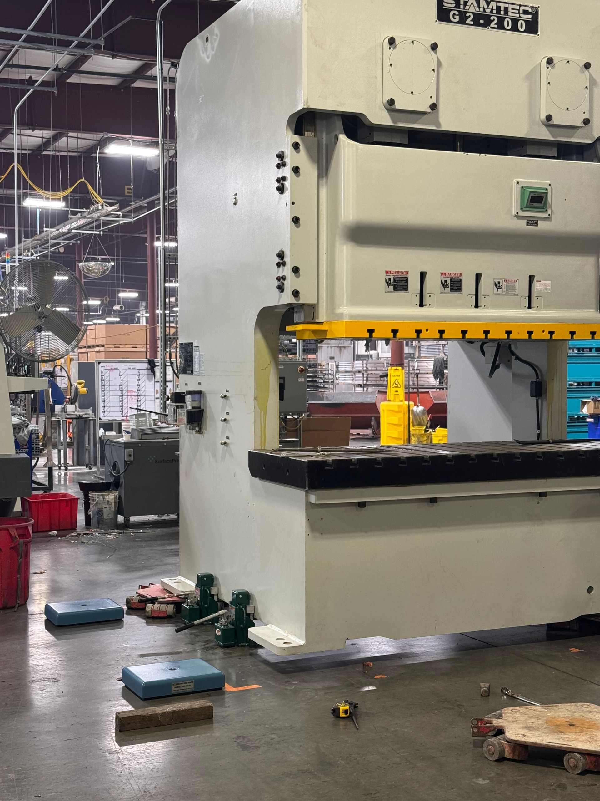 Large industrial stamping press in a factory setting, light gray, with a yellow safety barrier and debris on the floor.