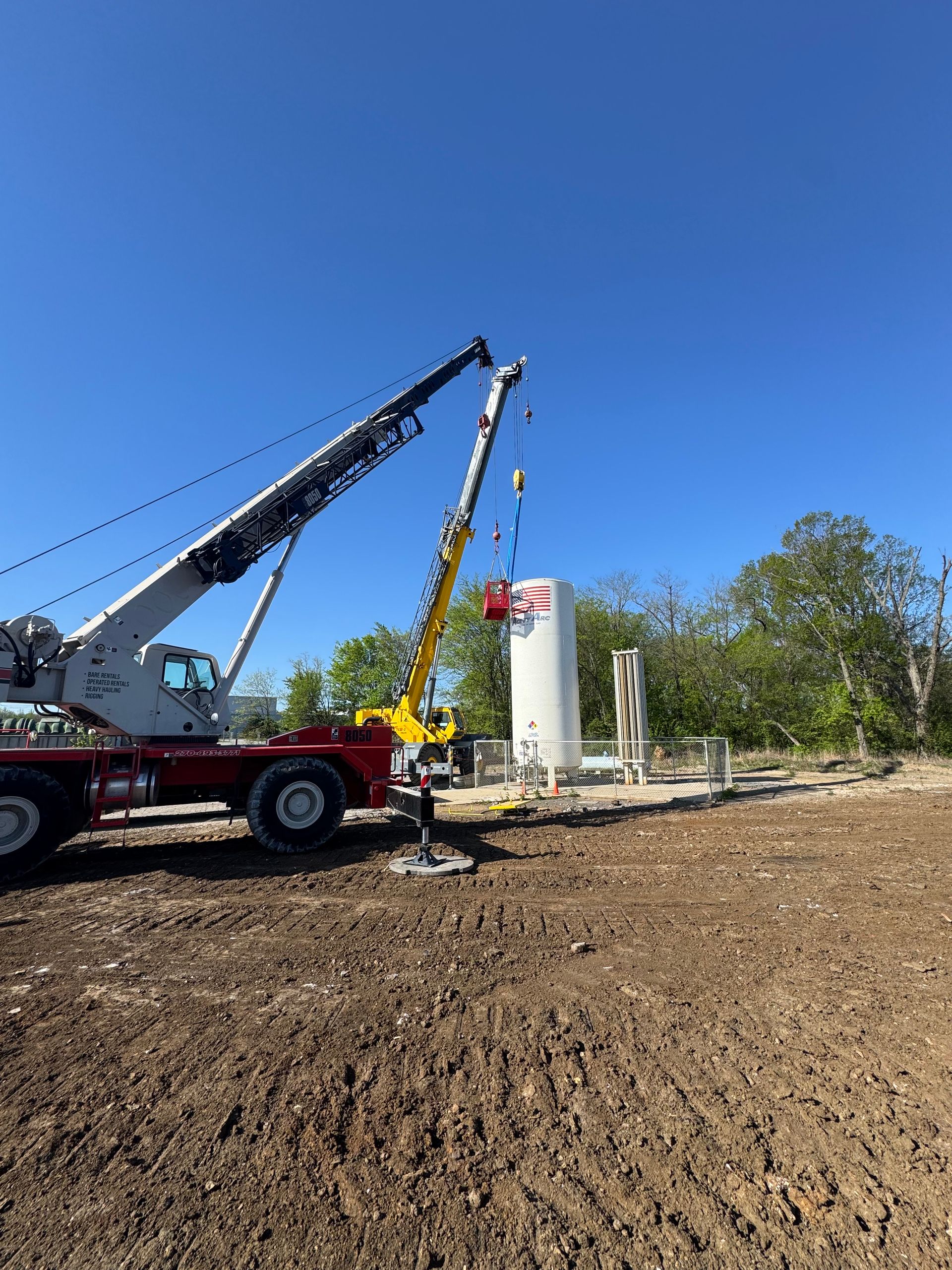 Two cranes lifting a large white tank outdoors against a blue sky.