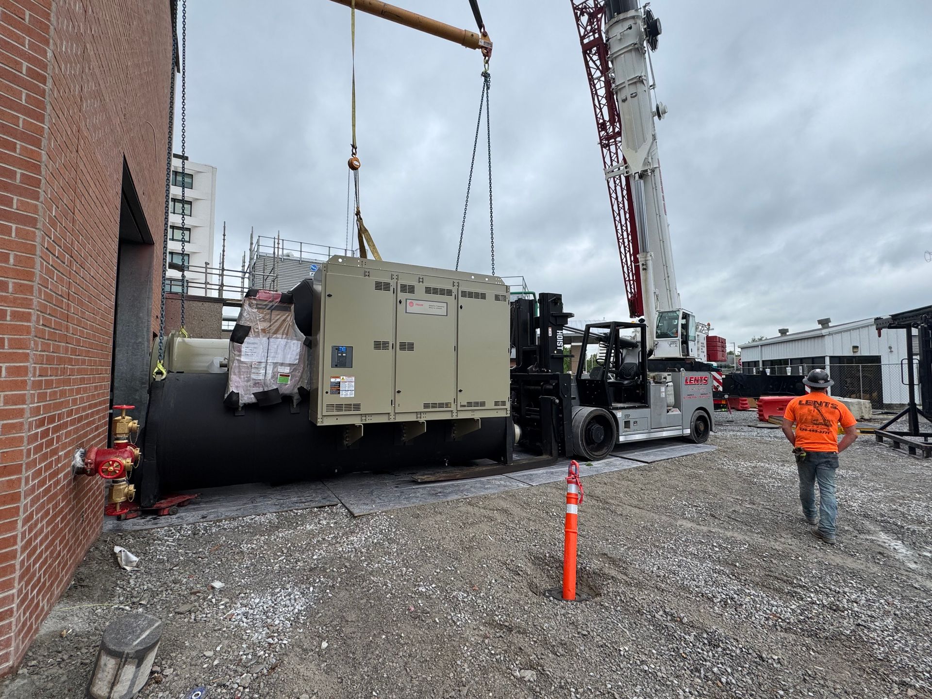 A crane lifting a generator and tank beside a brick building. Construction worker walks away.
