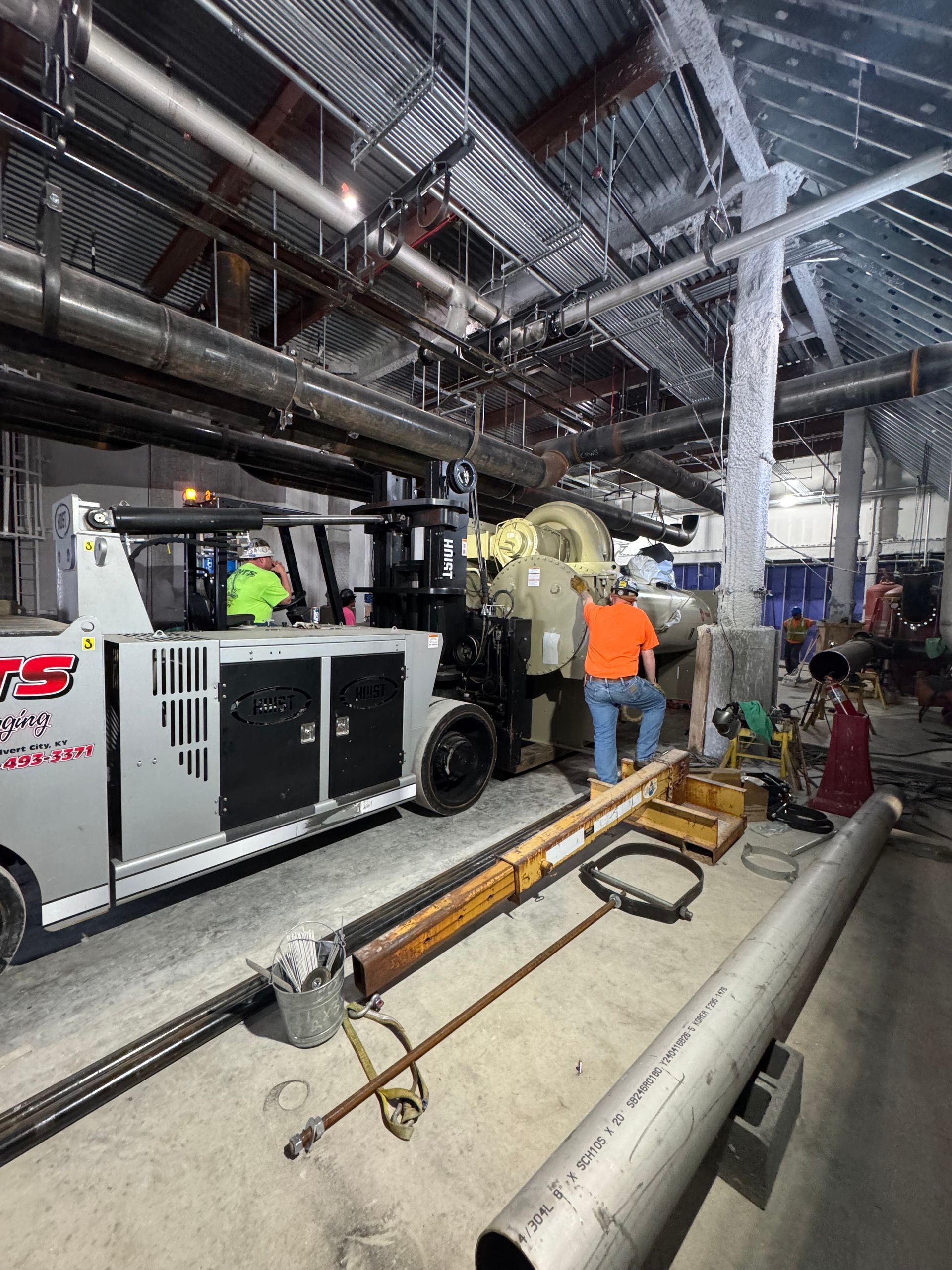 Workers installing equipment in a construction zone. Men in safety vests stand near machinery inside a building.