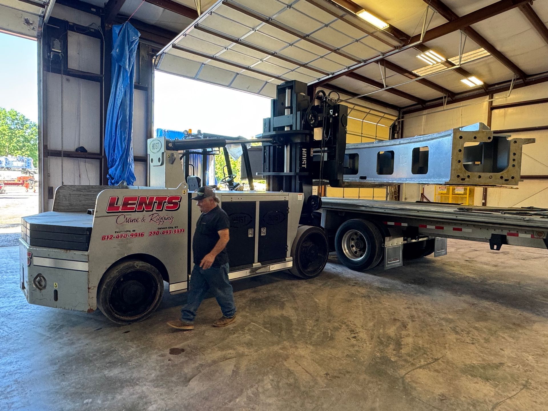 A man walks near a large industrial lifter carrying a metal structure inside a warehouse.