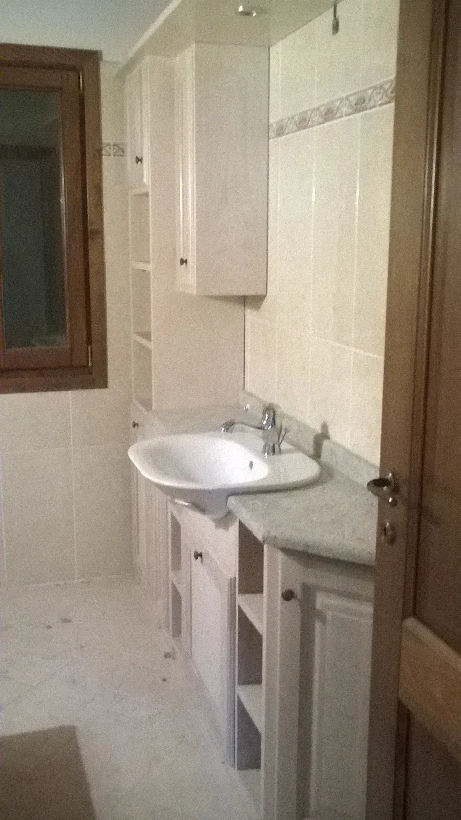 Bagno con piastrelle e mobili chiari, un lavandino angolare e una porta e una finestra con telaio in legno.