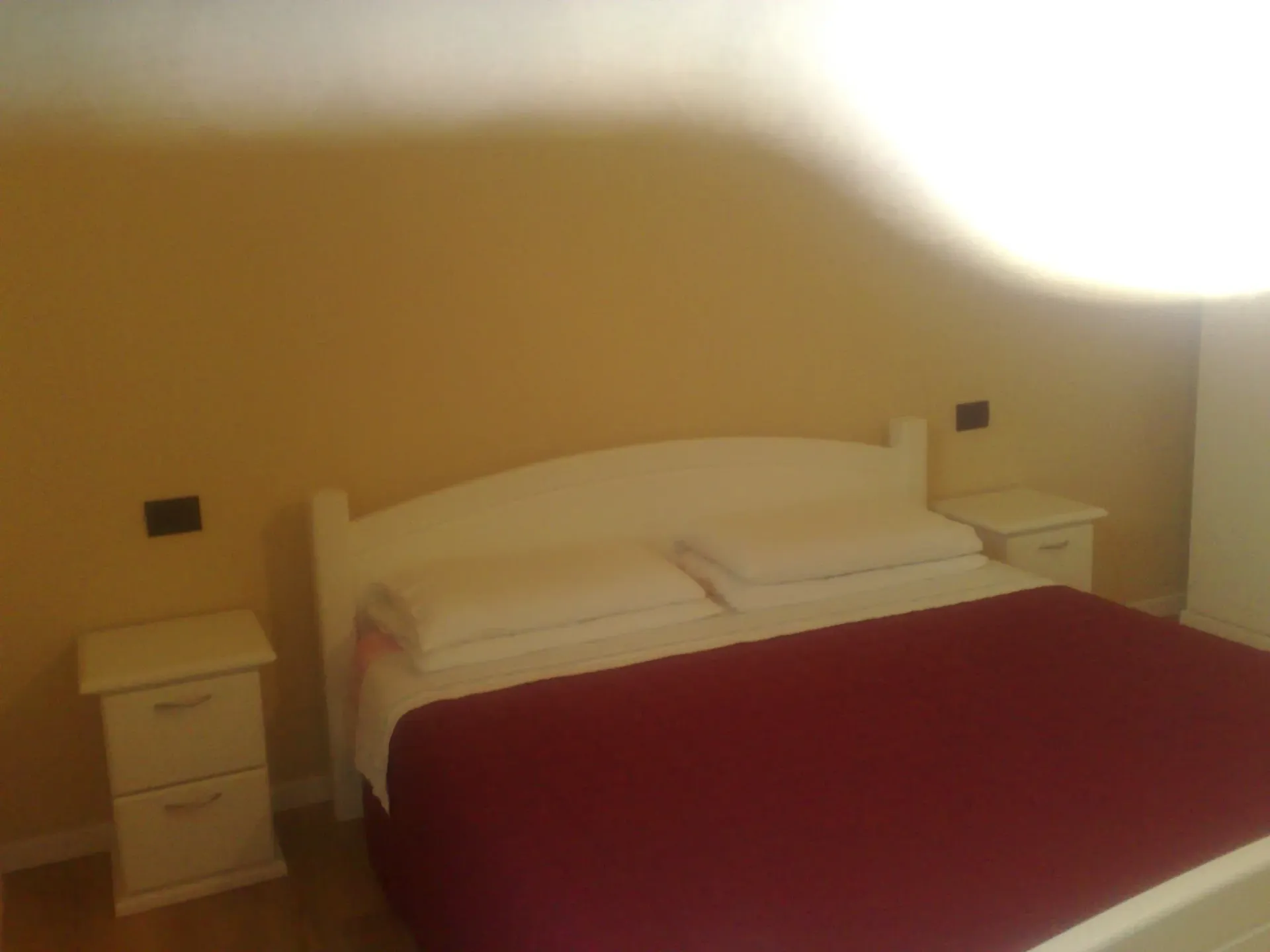 Camera da letto con letto bianco, comodini e copriletto bordeaux contro una parete gialla.