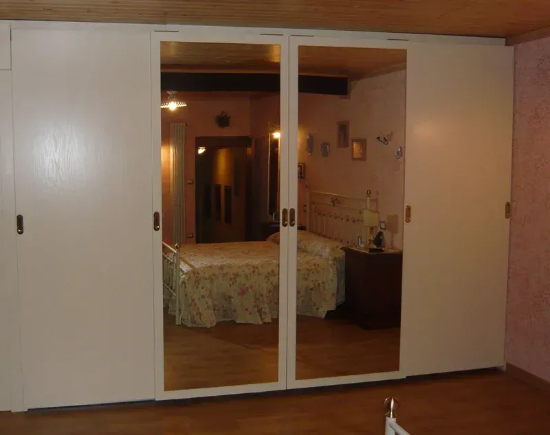 Le ante scorrevoli bianche dell'armadio rispecchiano una camera da letto con letto, comodino e decorazioni murali.