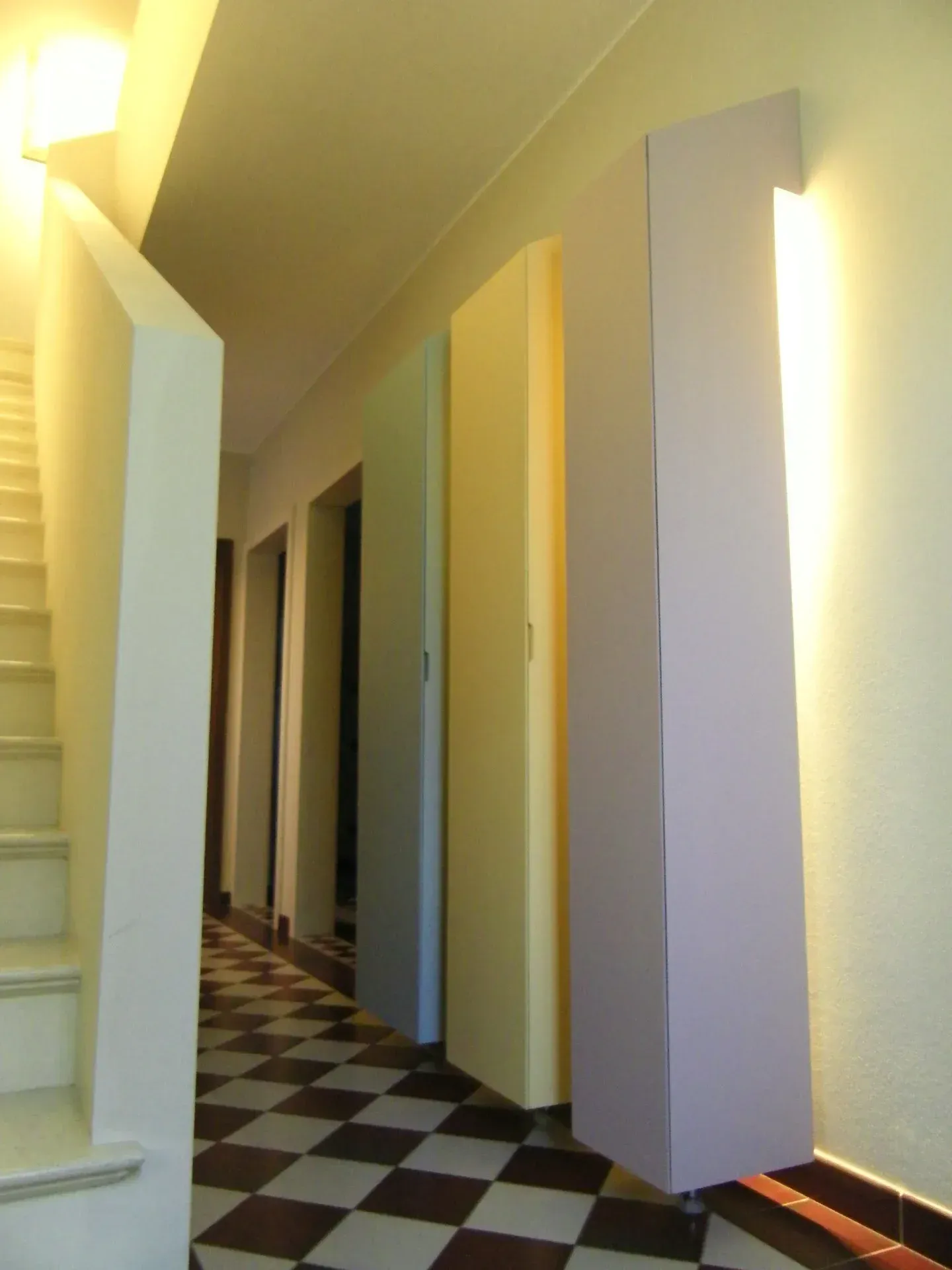 Corridoio con pavimento a scacchi, elementi rettangolari colorati alle pareti e scale sulla sinistra.