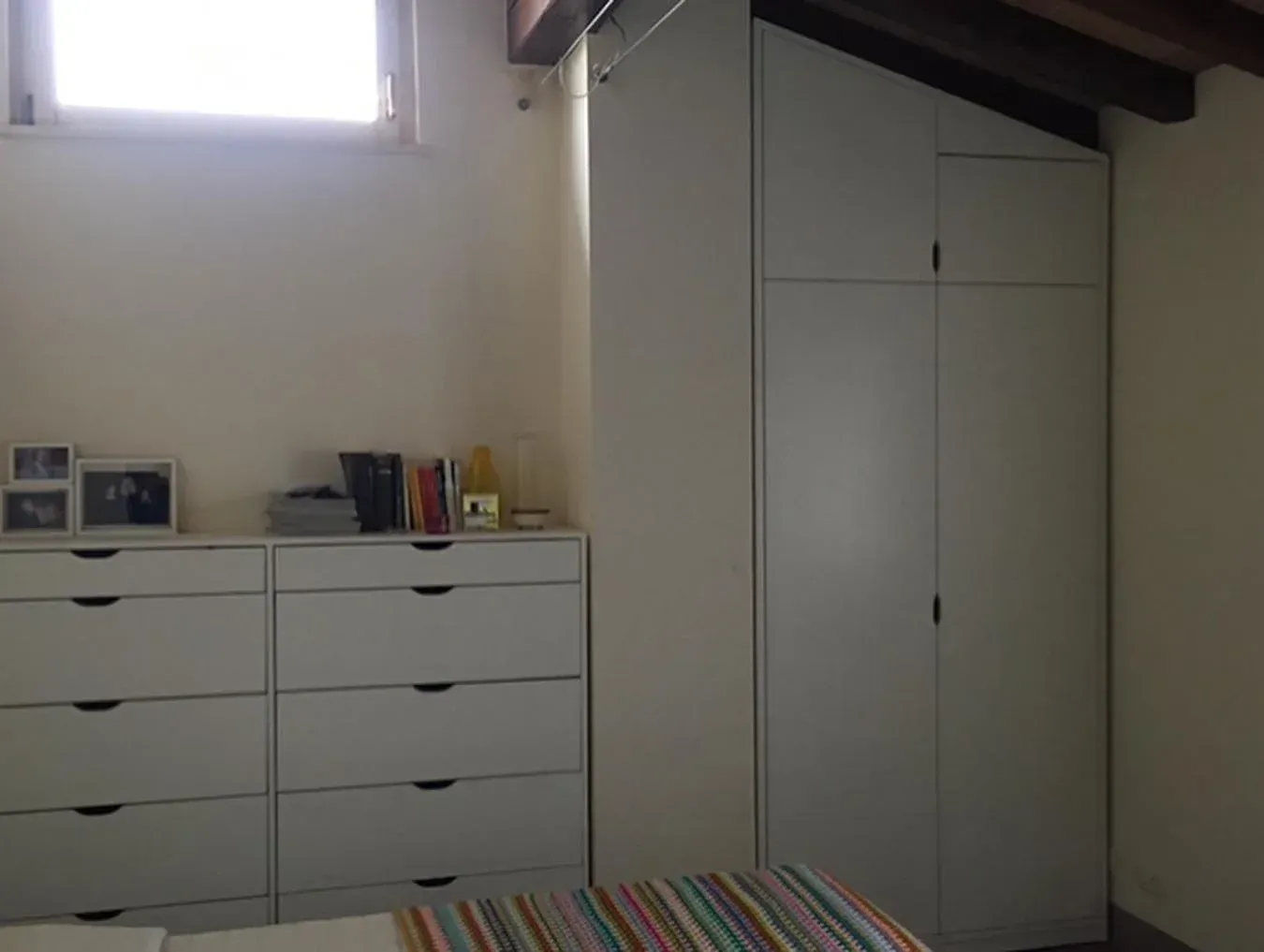 Camera da letto con cassettiere bianche, un armadio alto e una piccola finestra.