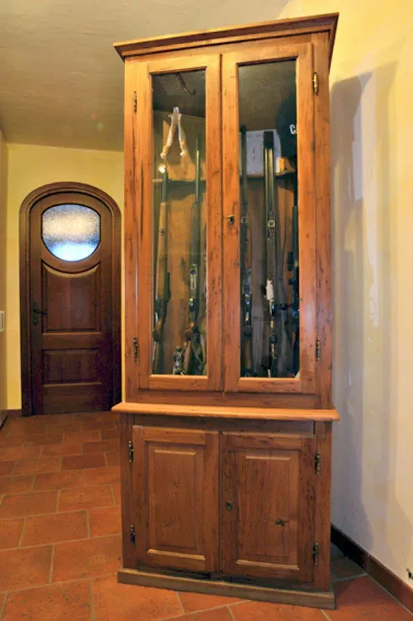 Mobiletto portafucili in legno con ante in vetro, contenente fucili. Legno marrone, su pavimento in cotto.