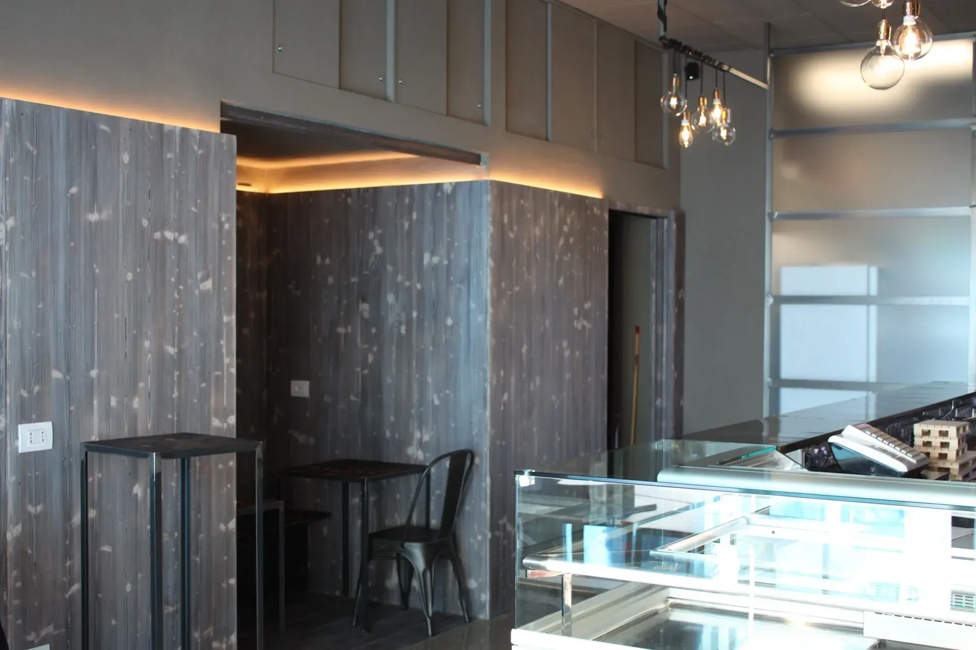Interno di un bar con pareti rivestite in legno, tavolino, vetrina in vetro e illuminazione da incasso.