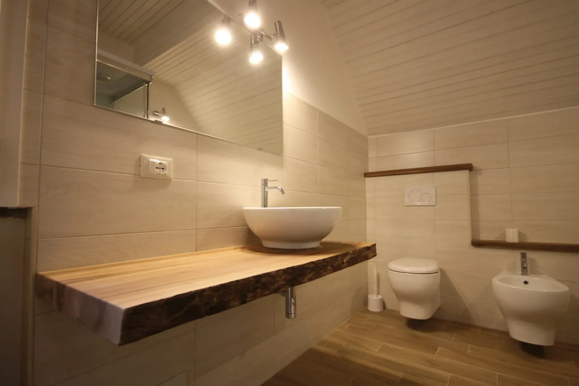 Bagno con piano di lavoro sospeso in legno, lavandino e WC bianchi; pareti piastrellate beige e pavimento in legno chiaro.