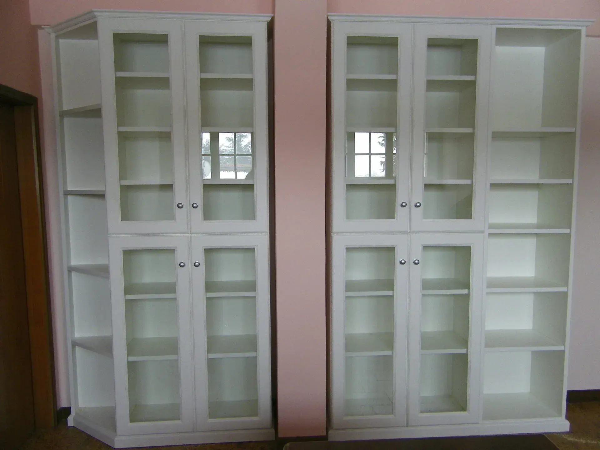 Due librerie bianche con ante in vetro e più ripiani contro una parete rosa. Una aperta.