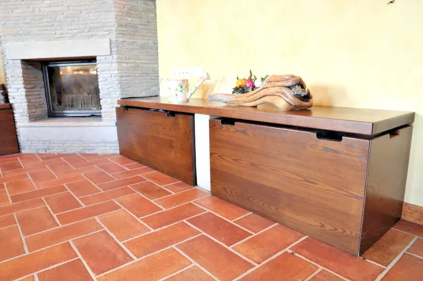 Panca portaoggetti in legno con oggetti sopra, appoggiata a una parete gialla e a un pavimento in mattoni.