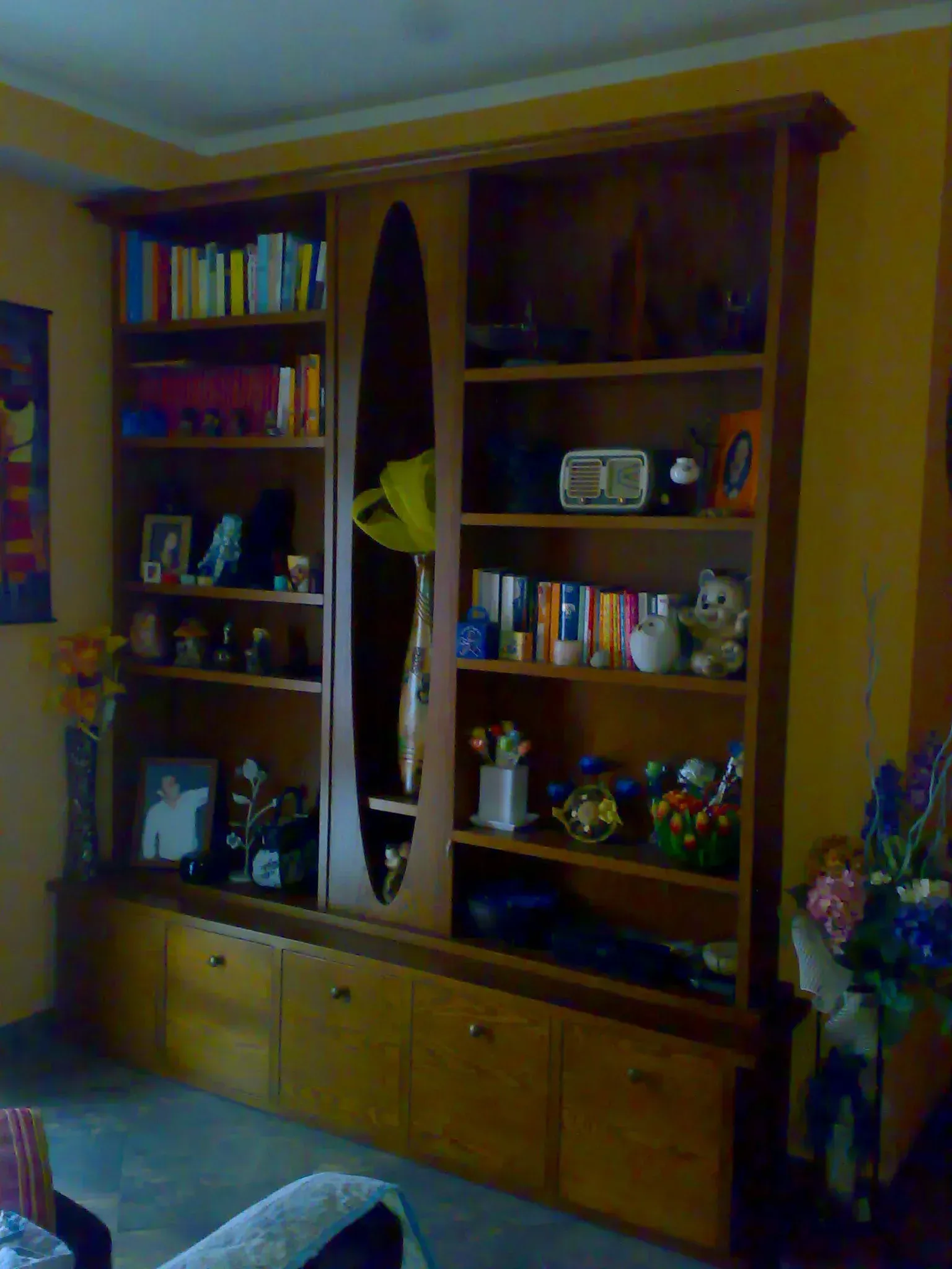 Libreria in legno piena di libri e oggetti decorativi, appoggiata a una parete gialla.