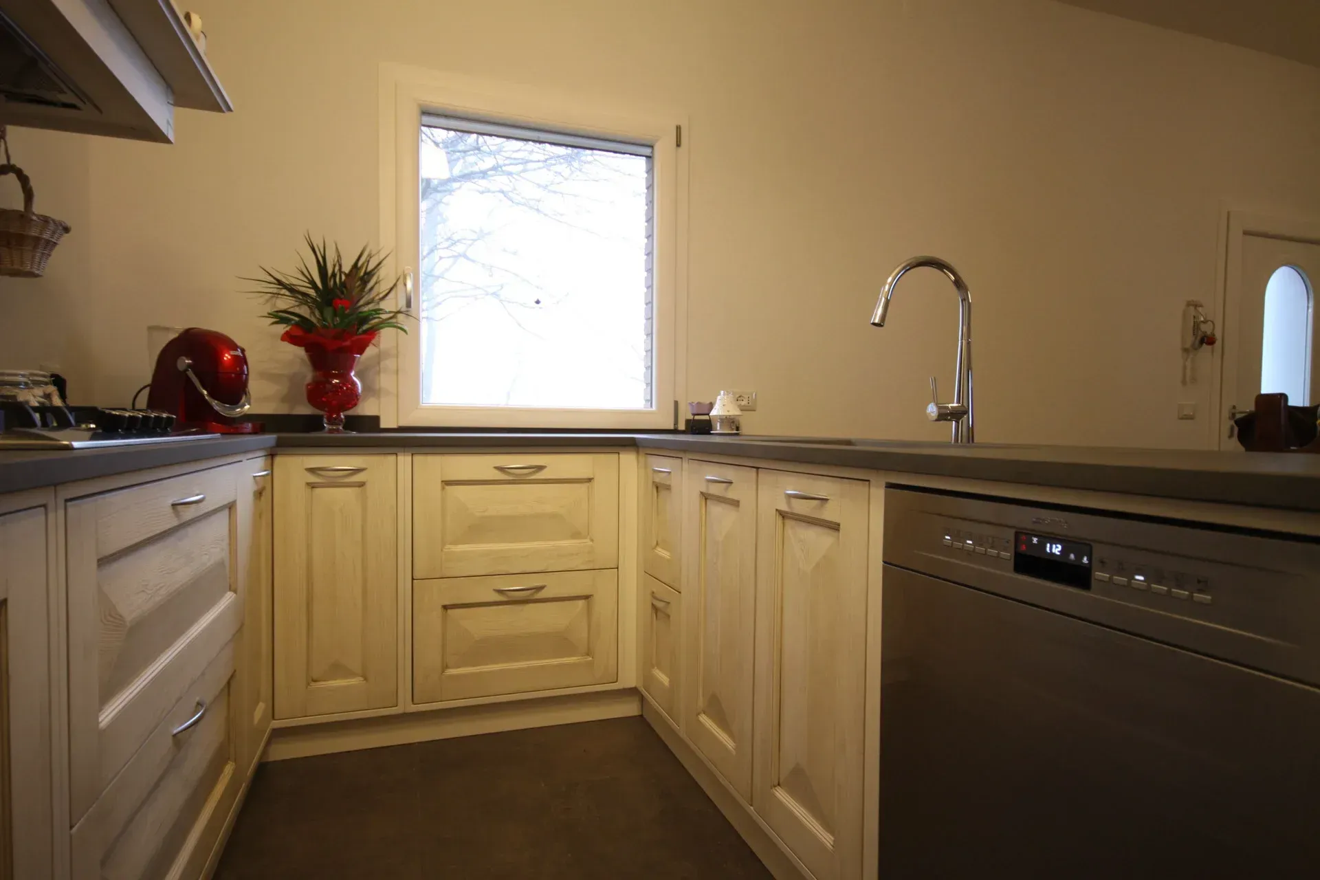 Mobili da cucina color crema con lavastoviglie in acciaio inox, finestra sopra il lavello e mixer rosso.