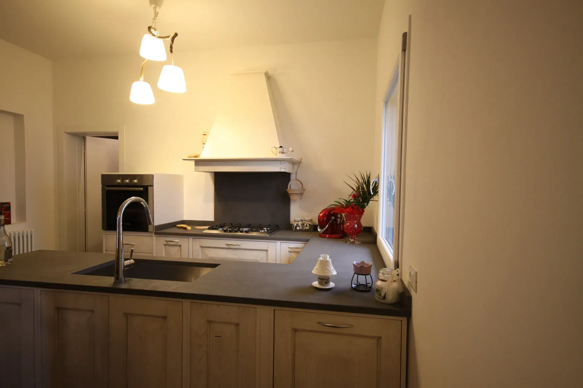 Cucina con isola, piano cottura, forno, cappa aspirante e lavello. Colori neutri, illuminazione da incasso.