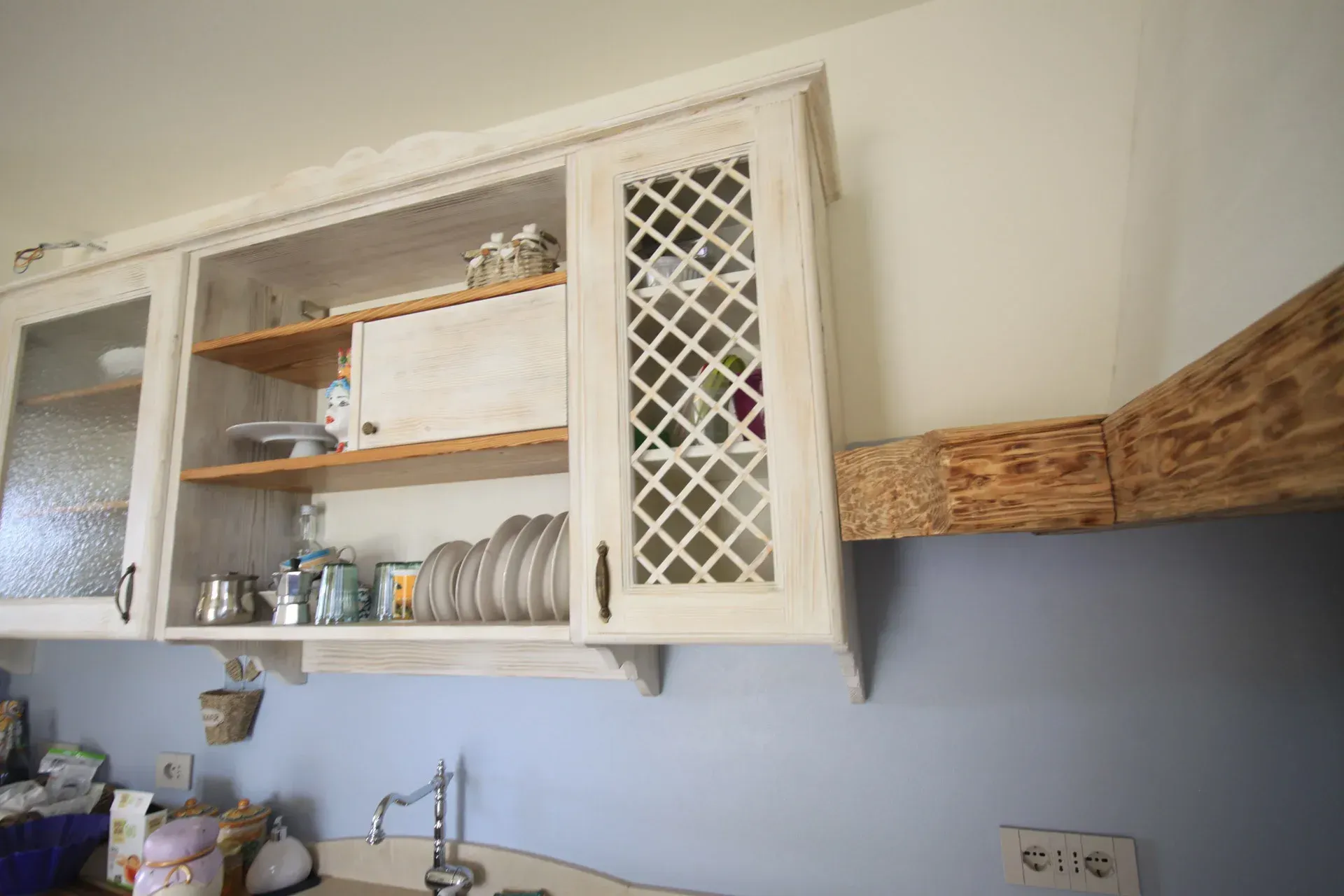 Mobili da cucina bianchi con ante in vetro e traliccio sopra un piano di lavoro. Trave in legno sulla destra.