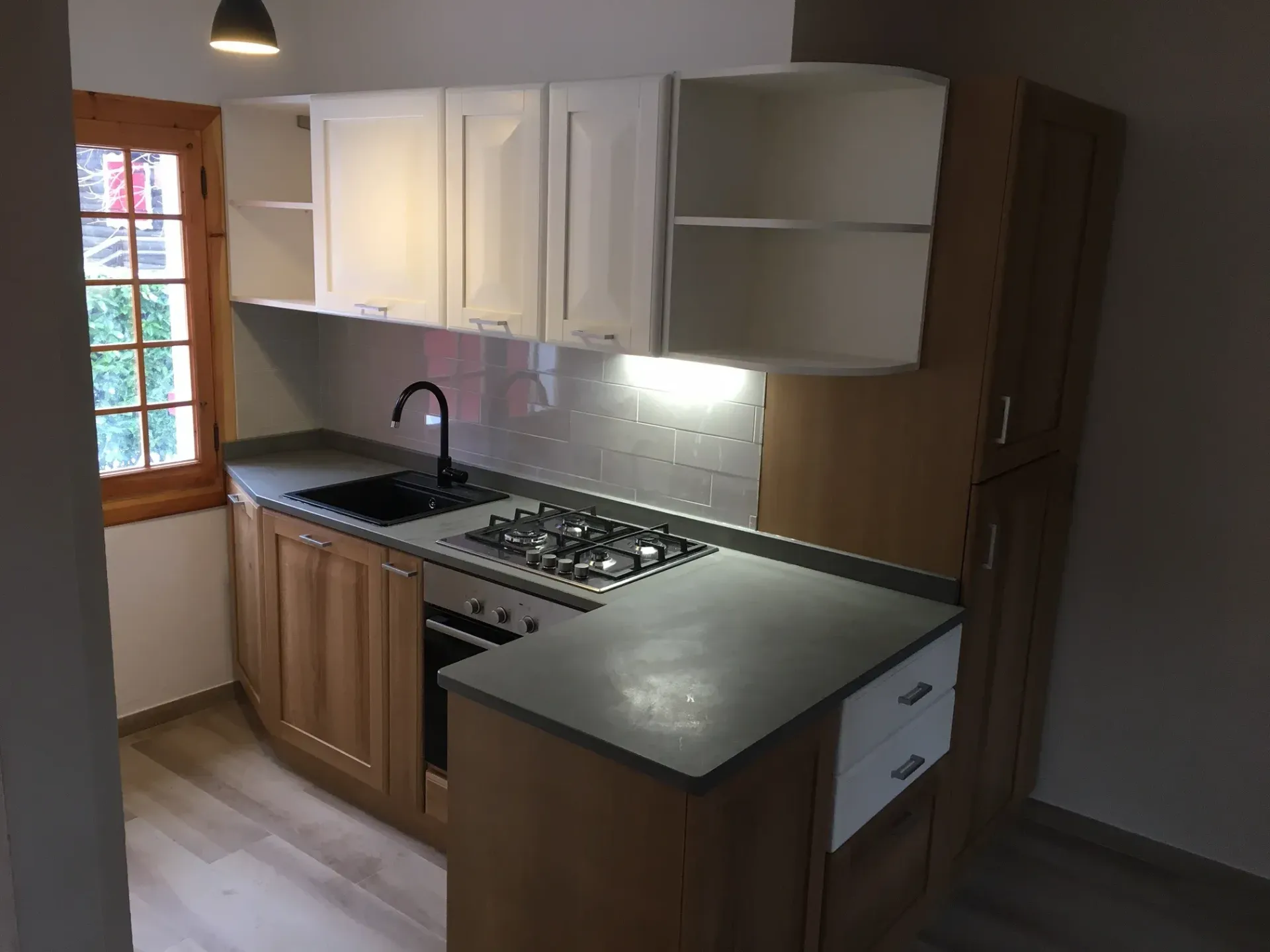 Piccola cucina con mobili bianchi e in legno, piano di lavoro grigio e piano cottura a gas.