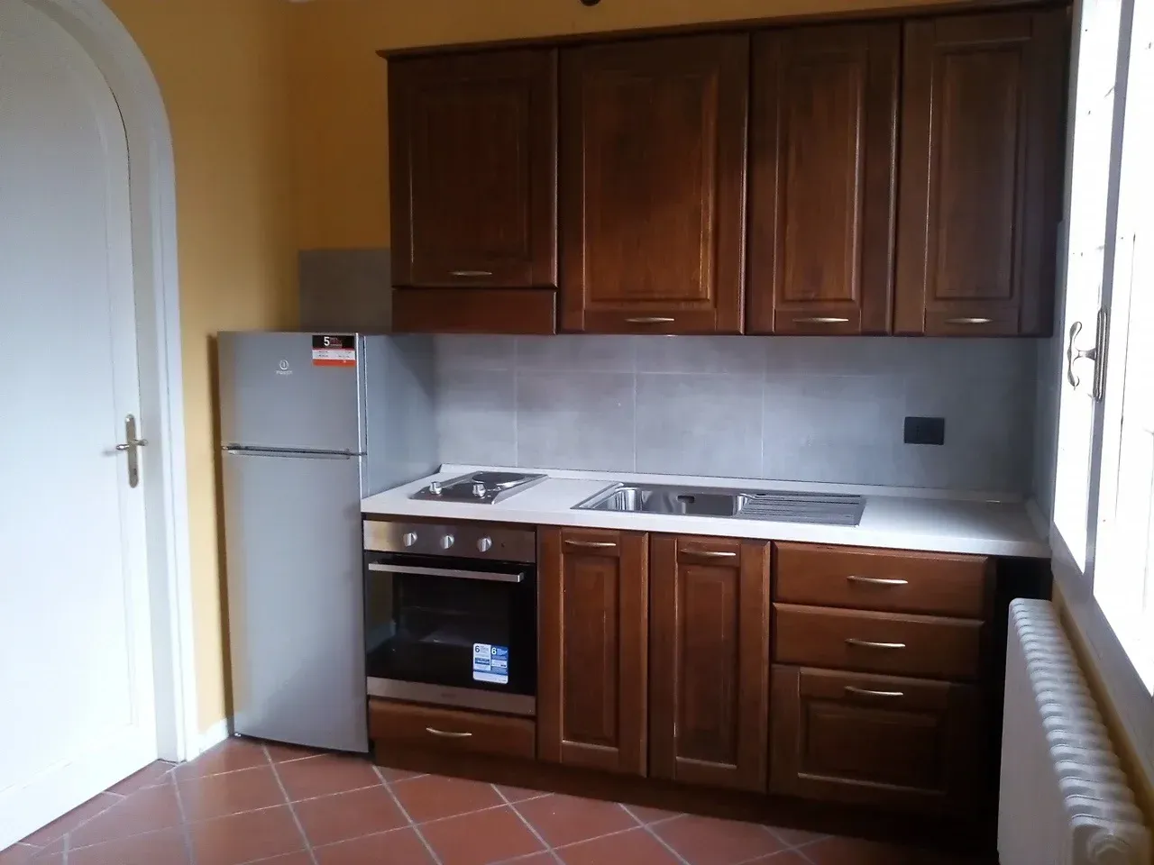 Piccola cucina con mobili marroni, frigorifero, forno e lavello, adagiata contro una parete gialla e pavimento piastrellato.