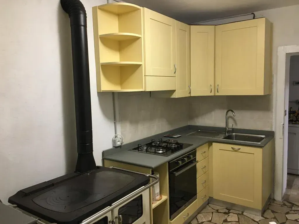 Cucina con mobili giallo chiaro, fornello nero, piano di lavoro grigio e tubo della stufa nero.