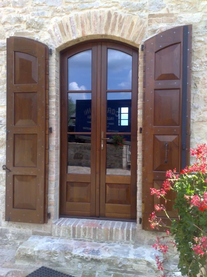 Porta in legno marrone con pannelli in vetro, fiancheggiata da persiane aperte, incastonata in un edificio in pietra.