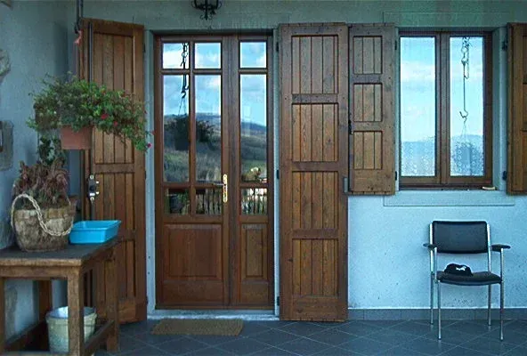 Porta e finestra in legno con persiane sul portico di un edificio azzurro, sedia e tavolo con piante.