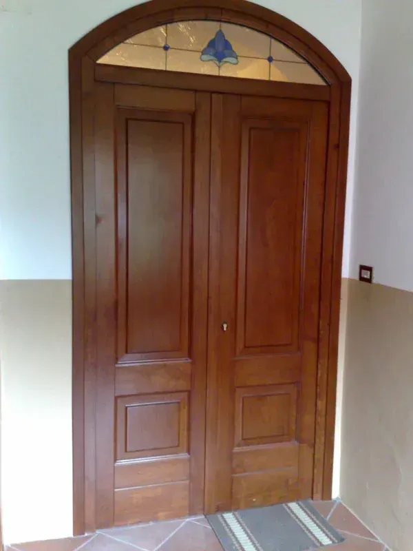 Doppia porta in legno con parte superiore arcuata e dettagli in vetro colorato.