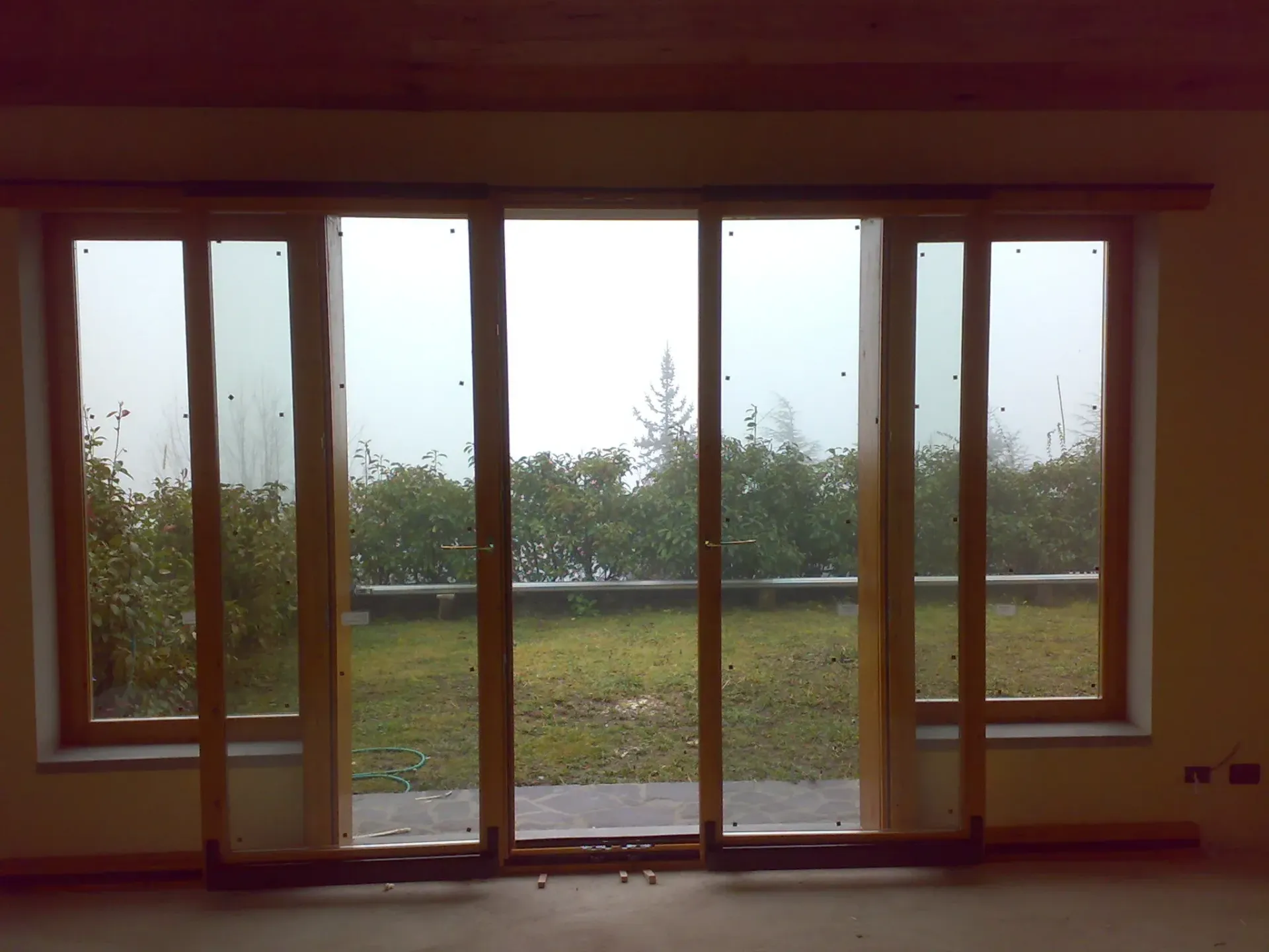 Porte scorrevoli in vetro con telai in legno si aprono su un cortile con cespugli e un cielo grigio.