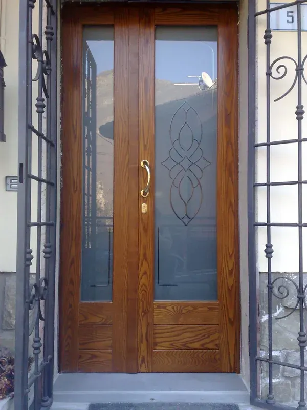 Doppie porte d'ingresso in legno con pannelli in vetro; una presenta un motivo decorato satinato, maniglia in metallo.