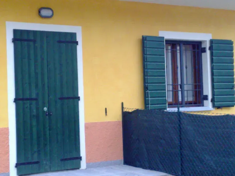 Porta e persiane verdi su un edificio giallo. Ferramenta nera, piccola finestra, recinzione nera.