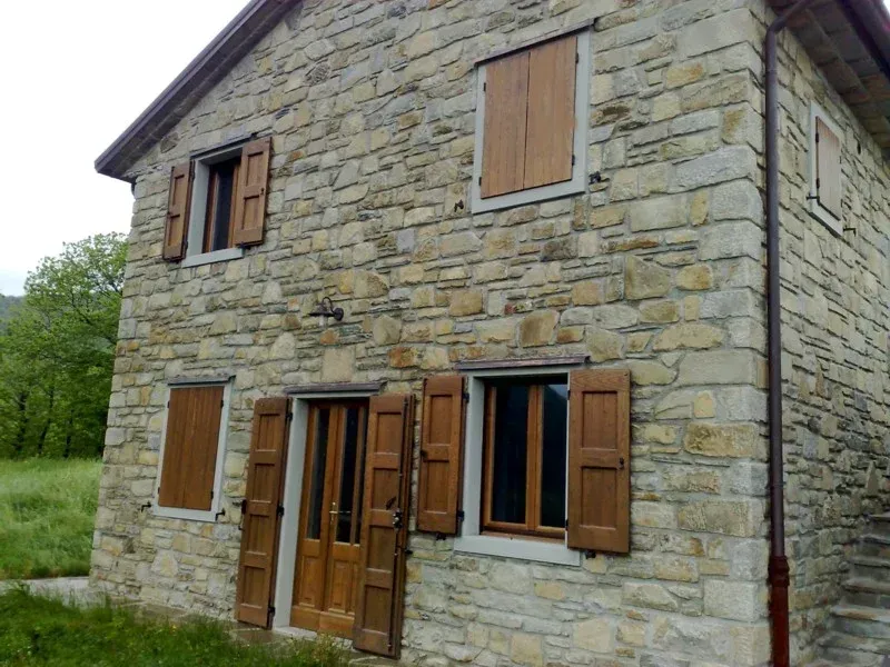 Casa in pietra con persiane e porta marroni. Erba verde e alberi sullo sfondo.