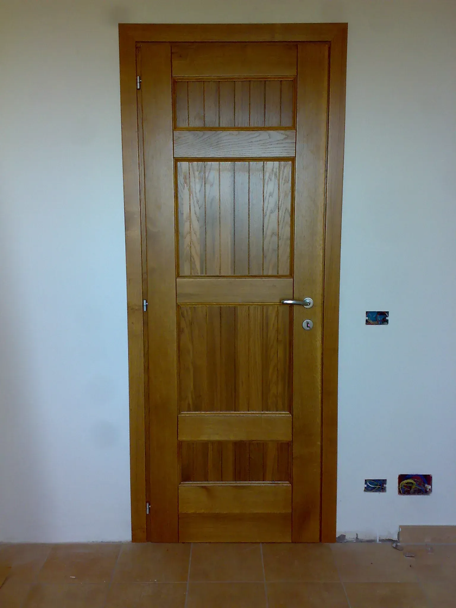 Porta in legno incastonata nella parete bianca; la porta ha un design a tre pannelli e una maniglia argentata.