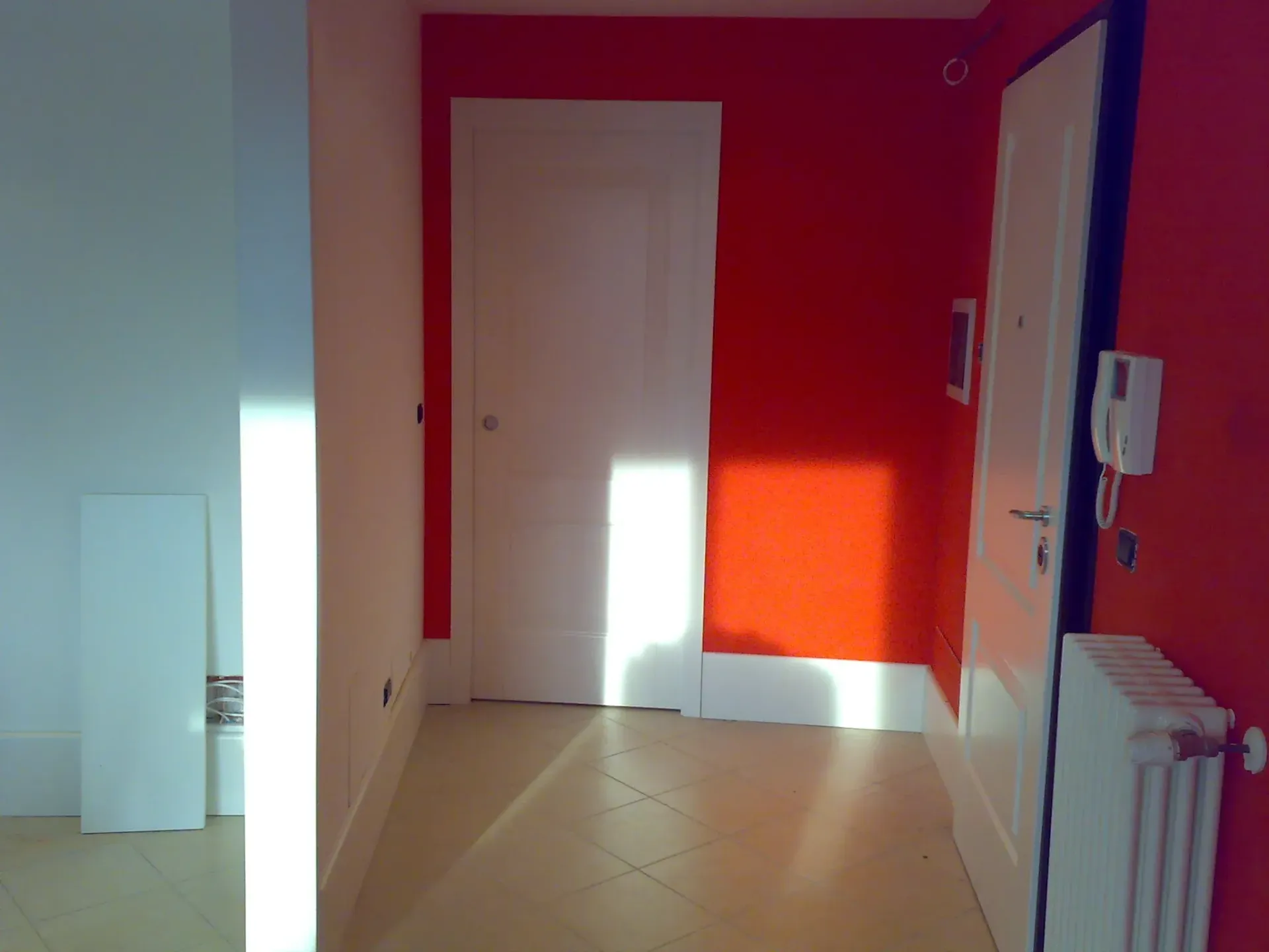 Corridoio con porte bianche, pareti rosse e un termosifone. La luce del sole filtra sul pavimento.