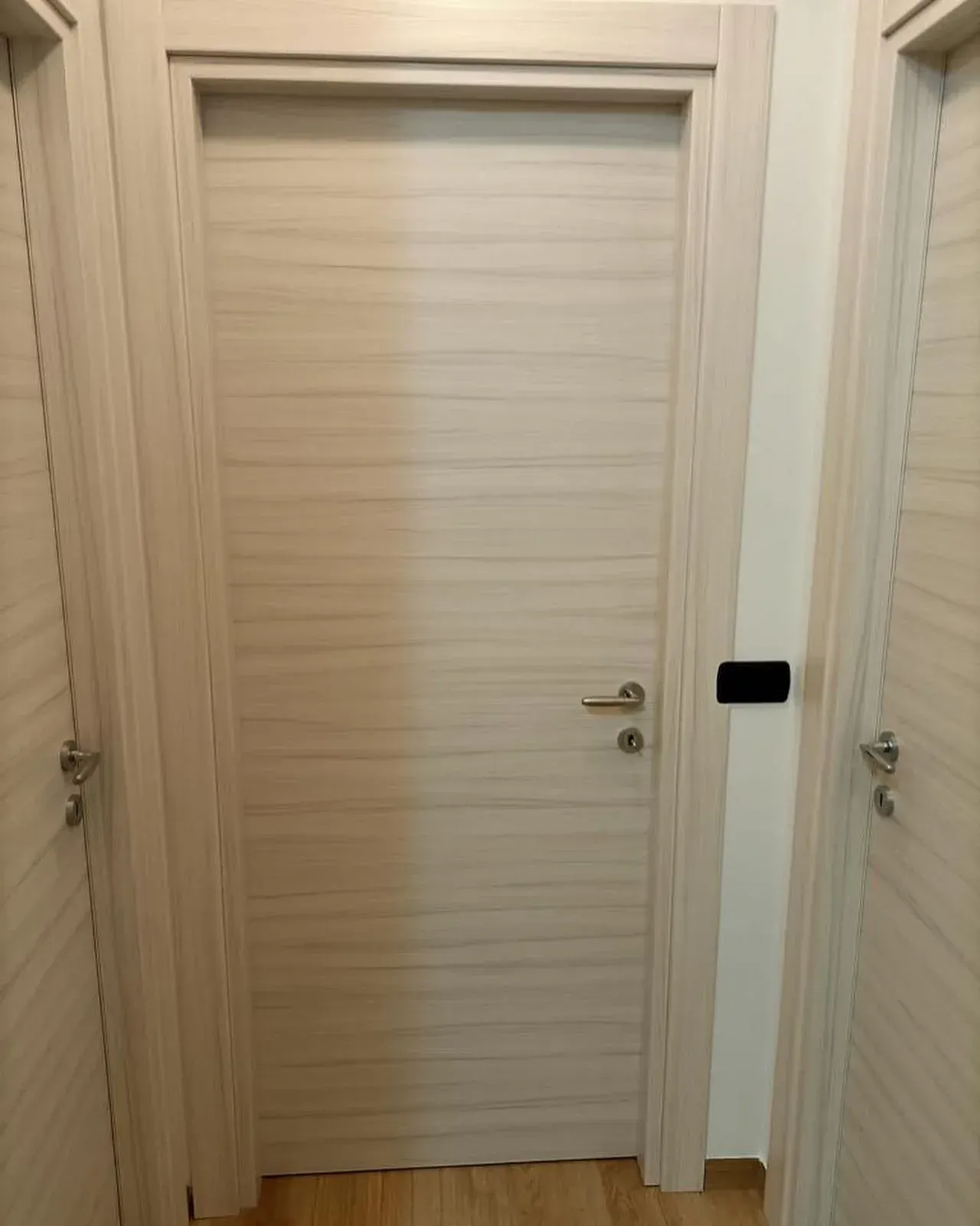 Porta beige in un corridoio, affiancata da porte simili. Include maniglia e interruttore nero.