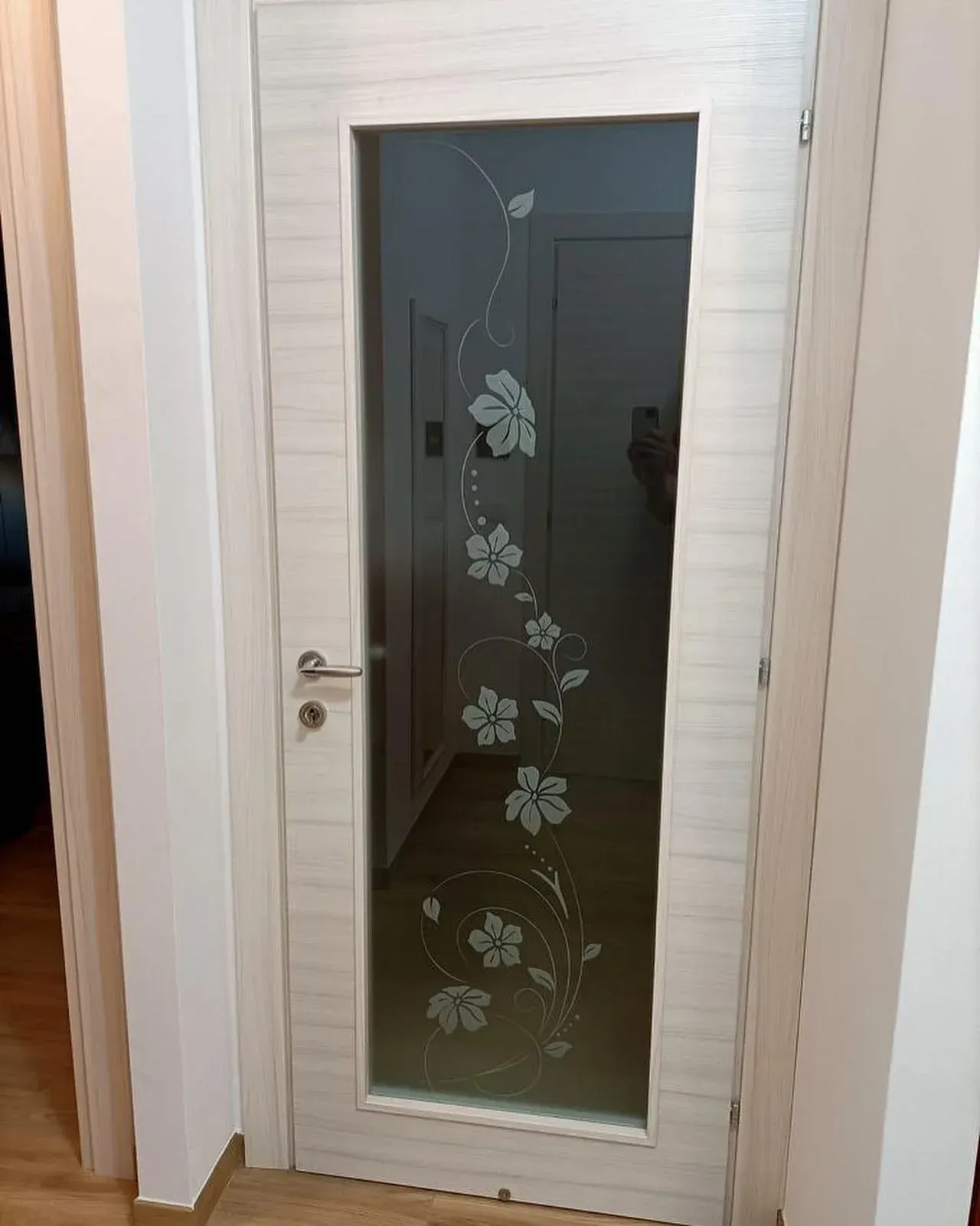 Porta bianca con pannello in vetro smerigliato e motivo floreale, che conduce a un corridoio.