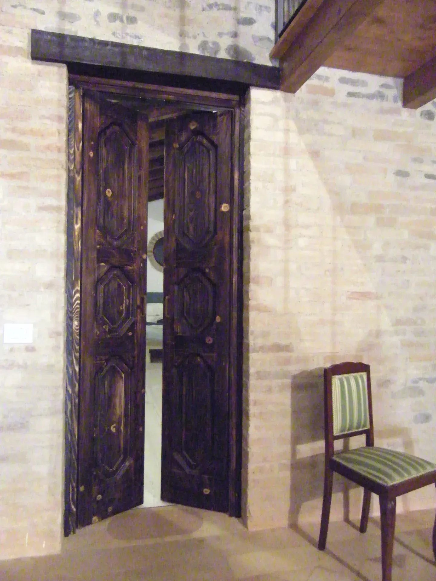 Doppie porte di legno scuro si aprono su una stanza. Una sedia è appoggiata sulla destra. Le pareti sono in mattoni chiari.