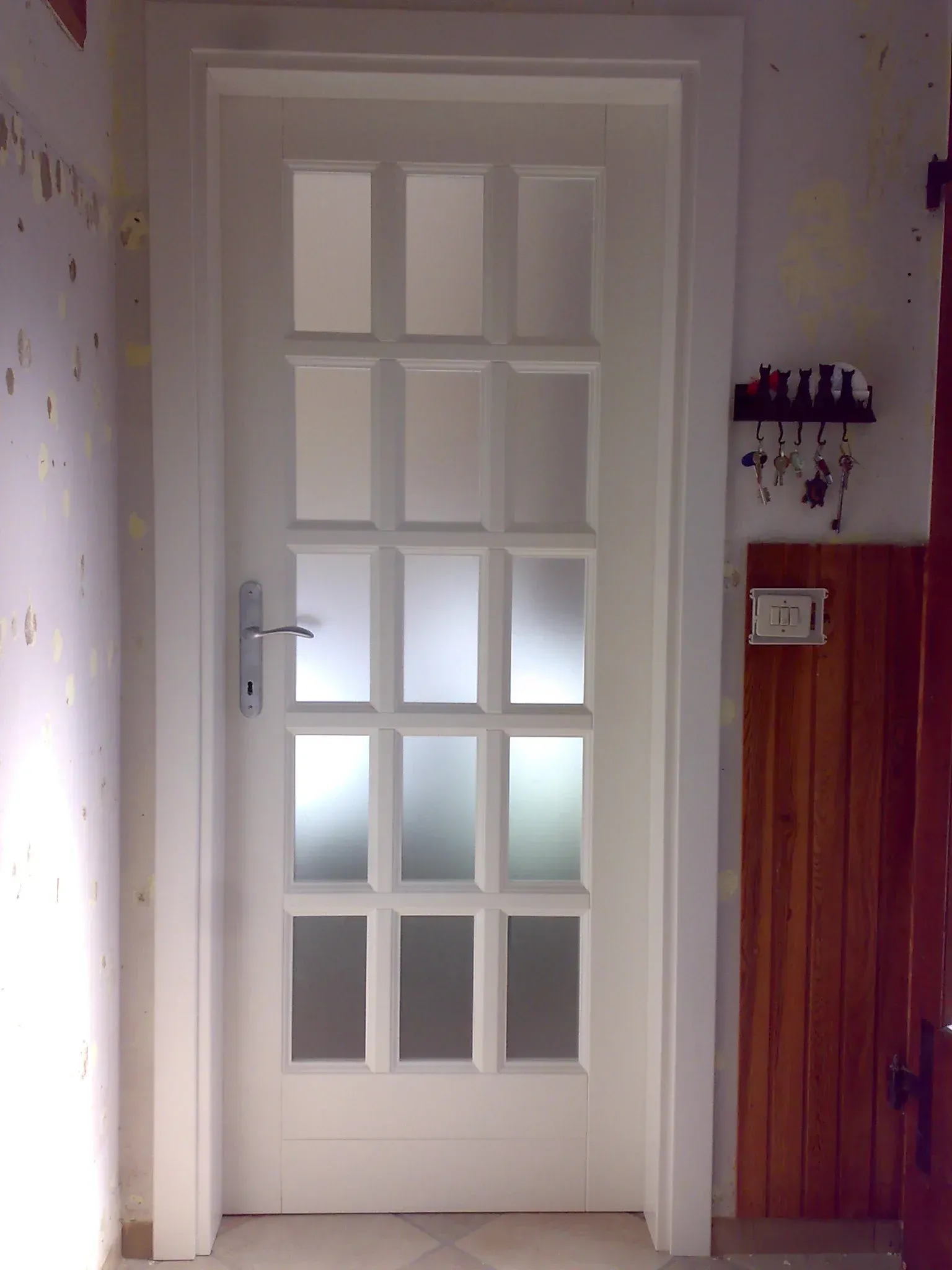 Porta bianca con vetri. Maniglia argentata. Portachiavi e pannello in legno a destra.