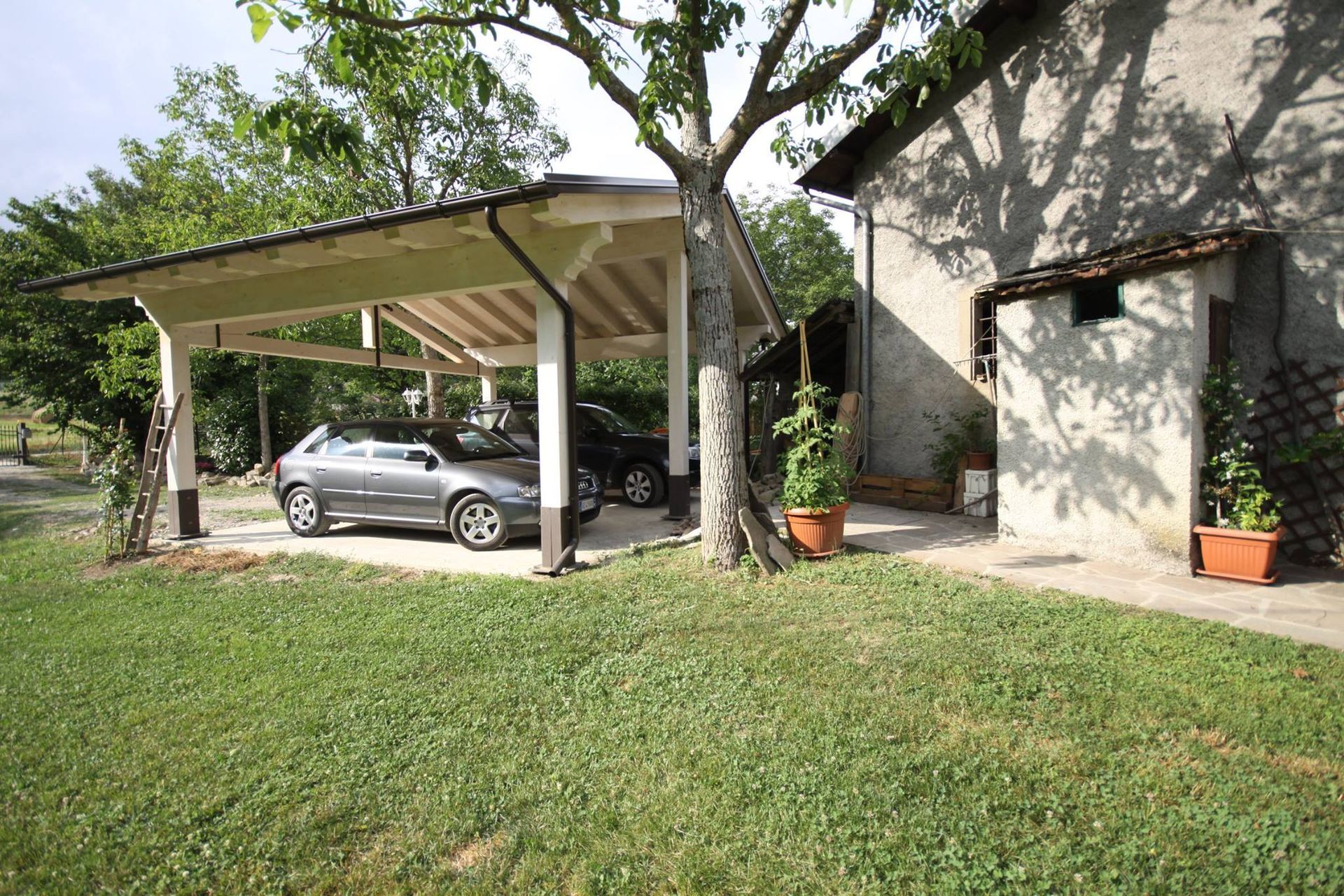 Auto parcheggiate sotto un posto auto coperto attaccato a un edificio in pietra, albero di fronte, cortile erboso.