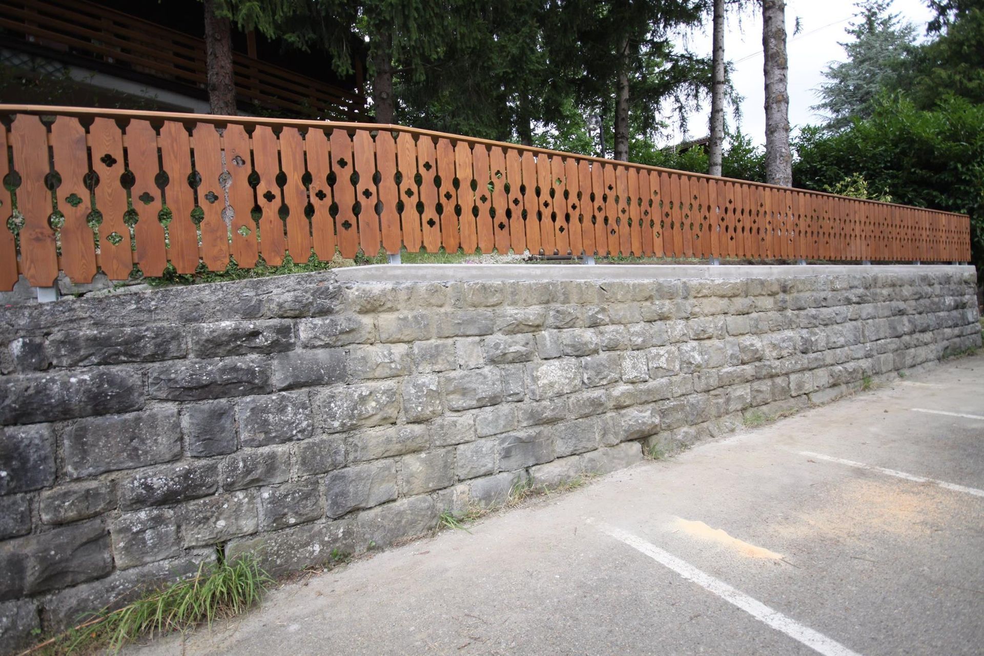 Muro di contenimento in pietra con recinzione decorativa marrone sopra, accanto all'asfalto.