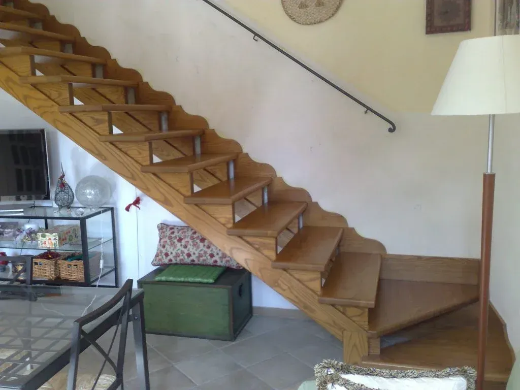 Scala in legno con bordo decorativo, corrimano su parete bianca, lampada e mobili.