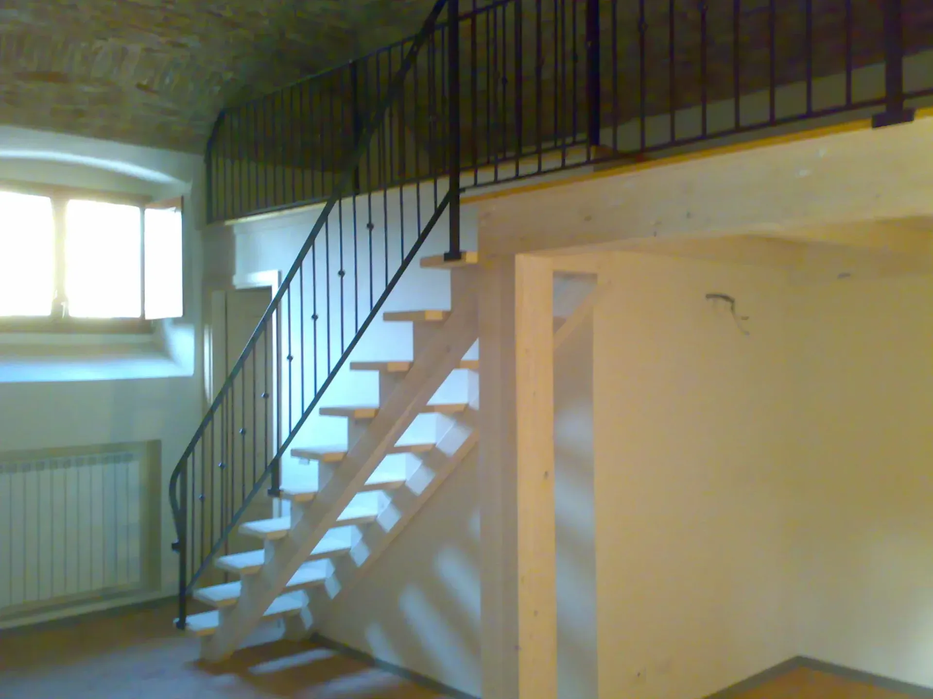Vista interna di un edificio con scala che conduce al soppalco. Pareti bianche, soffitto in mattoni, ringhiera nera.