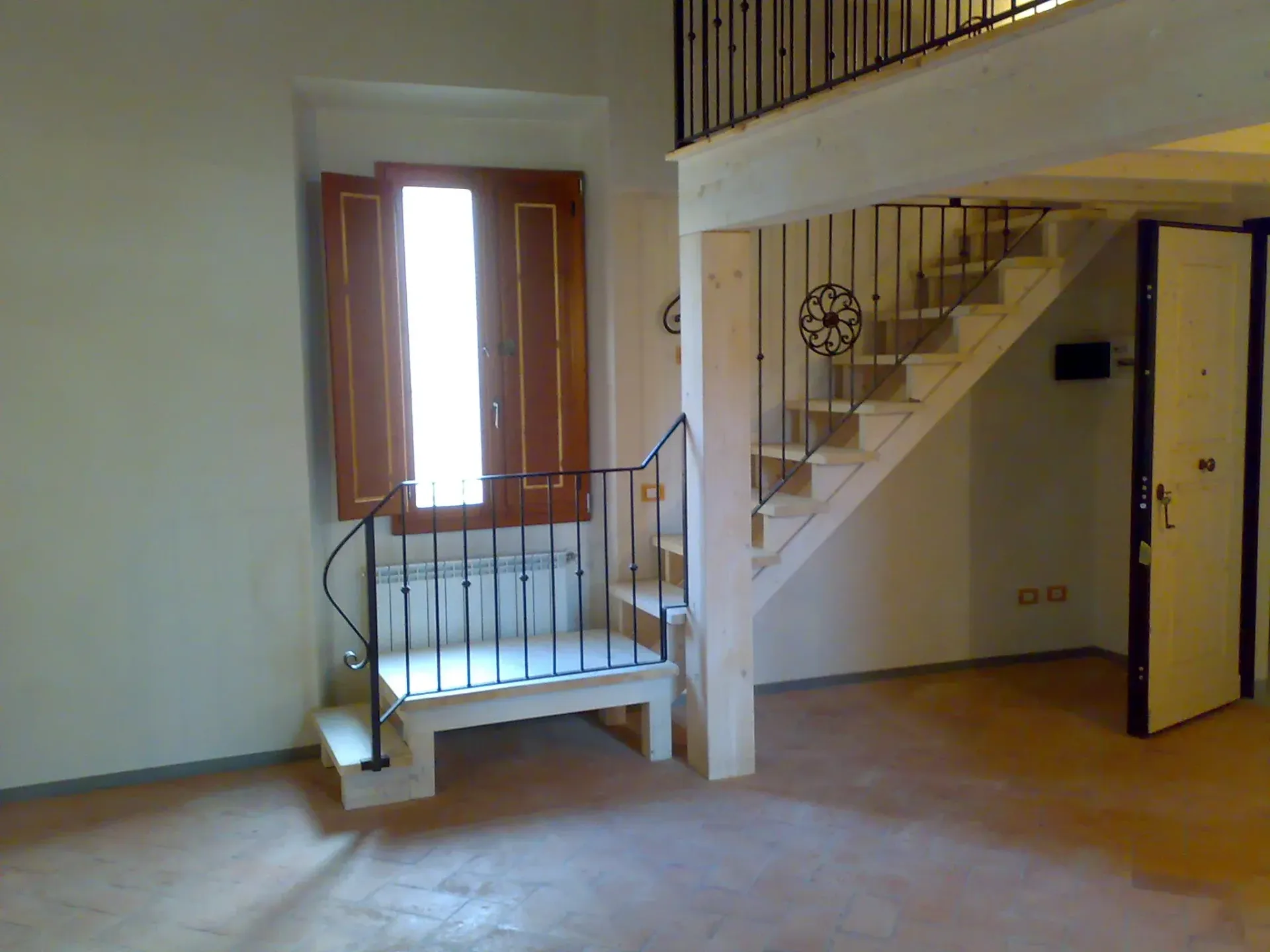 Interno con scala, finestra con persiane e piano superiore. Toni marroni e bianchi in tutto l'appartamento.