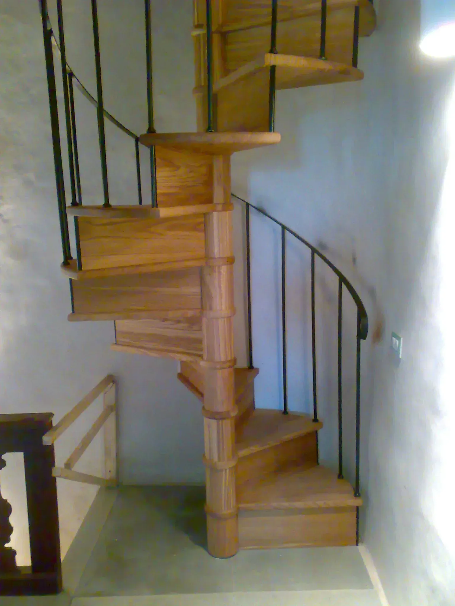 Scala a chiocciola in legno con ringhiere in metallo nero contro una parete di colore chiaro.