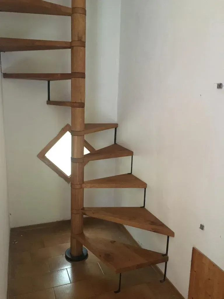Scala a chiocciola in legno con palo centrale, posta in un angolo con pareti bianche.