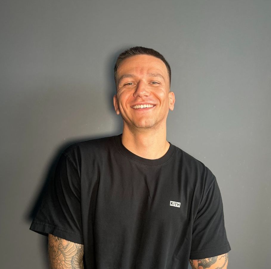 A man with tattoos is wearing a black t-shirt and smiling.