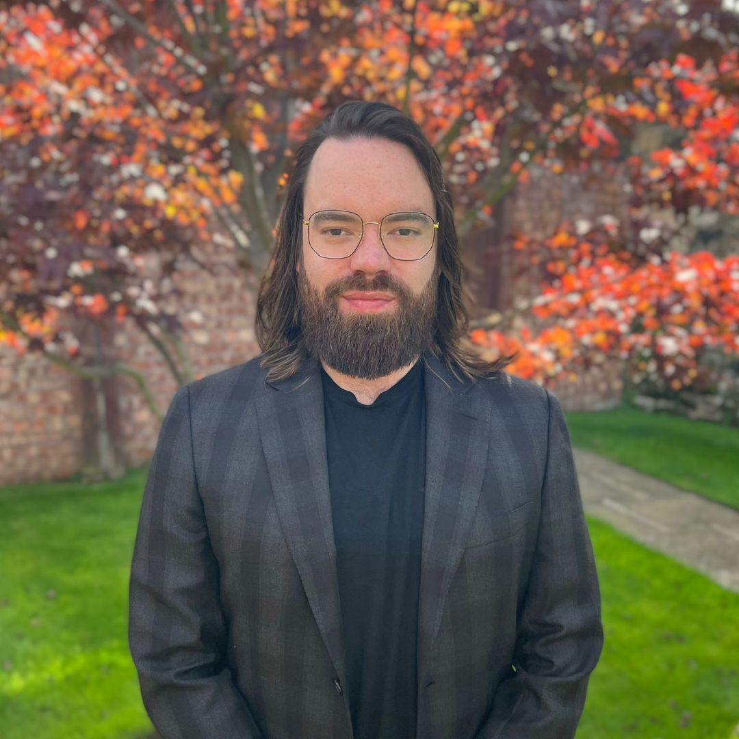 A man with a beard and glasses is standing in front of a tree.
