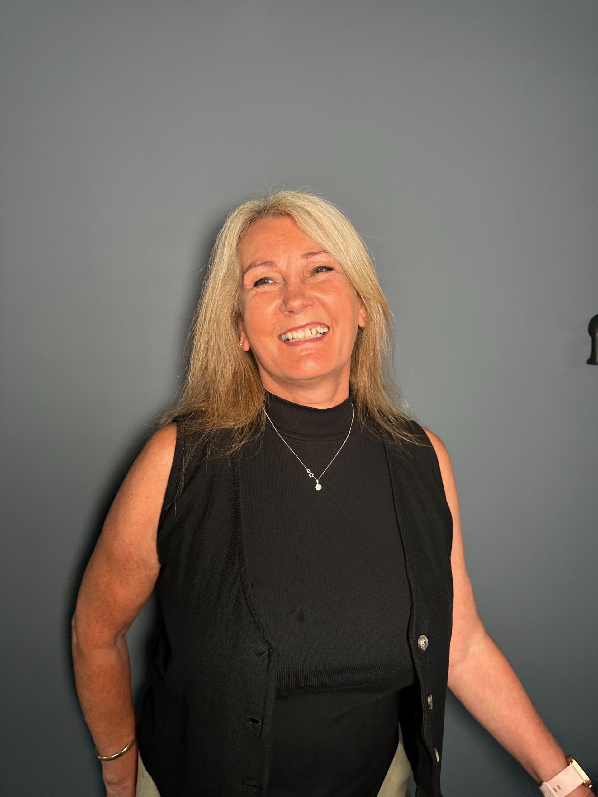 A woman wearing a black vest and a necklace smiles for the camera