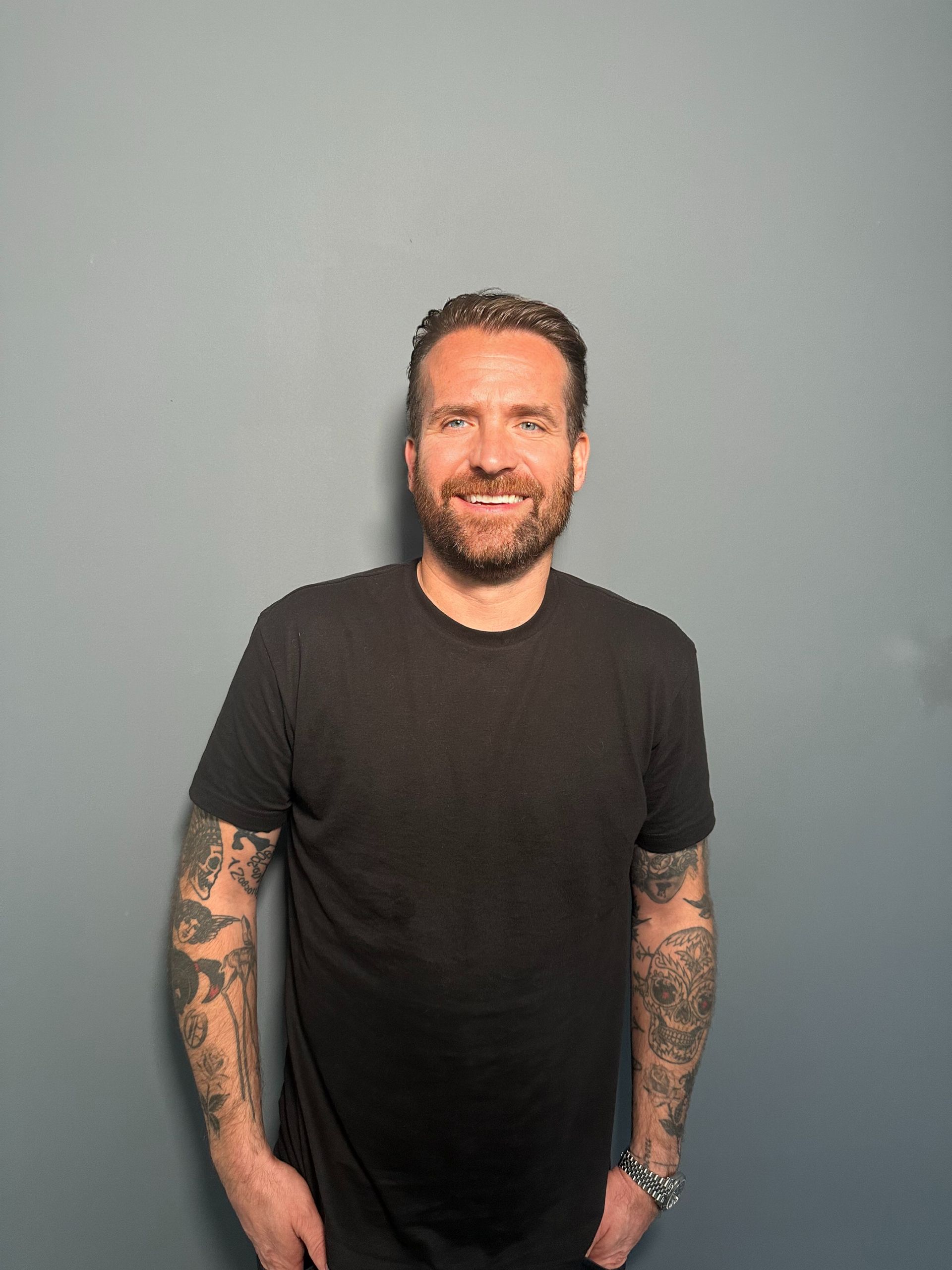 A man with a beard and tattoos is standing in front of a blue wall.