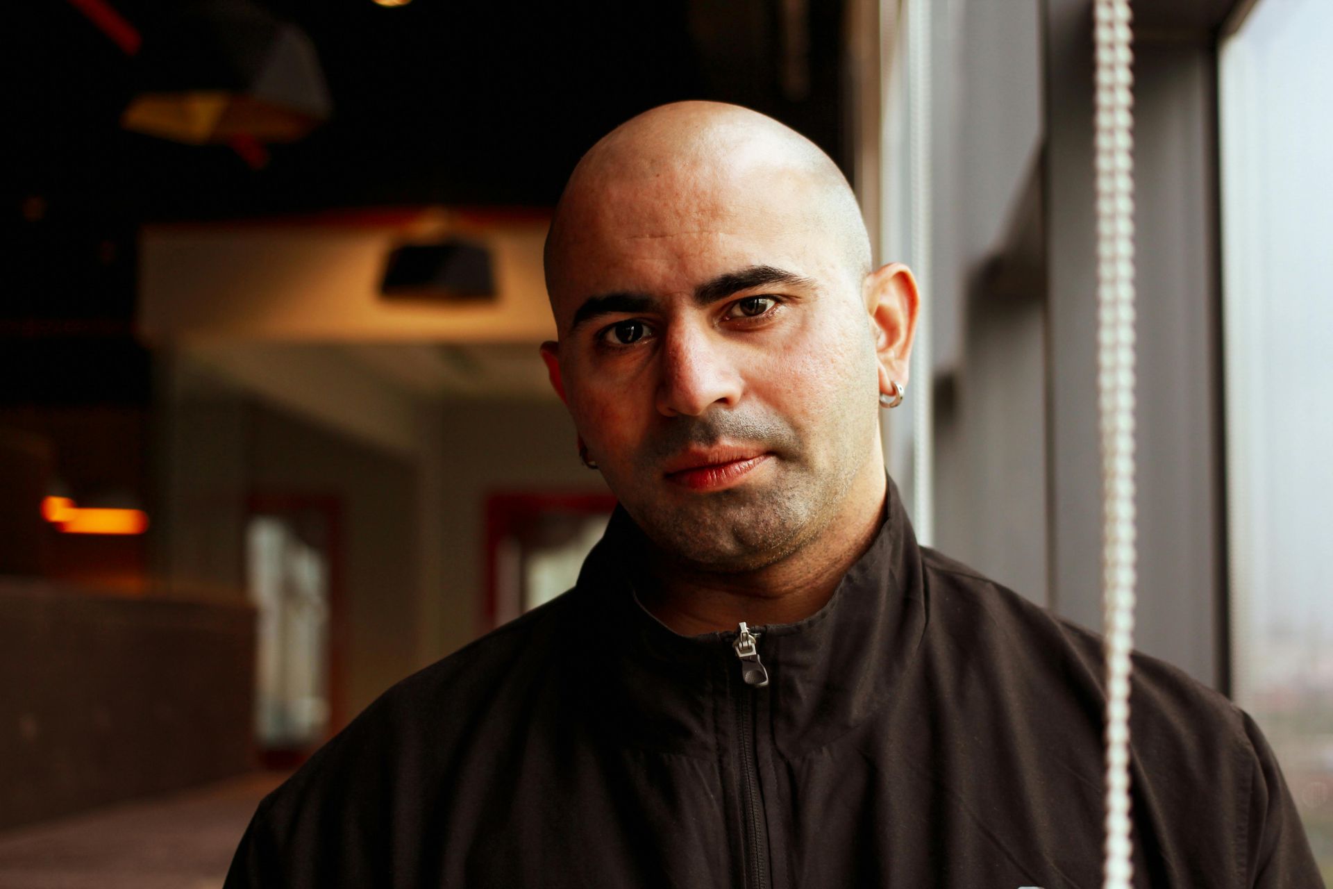 A bald man in a black jacket is standing in front of a window.